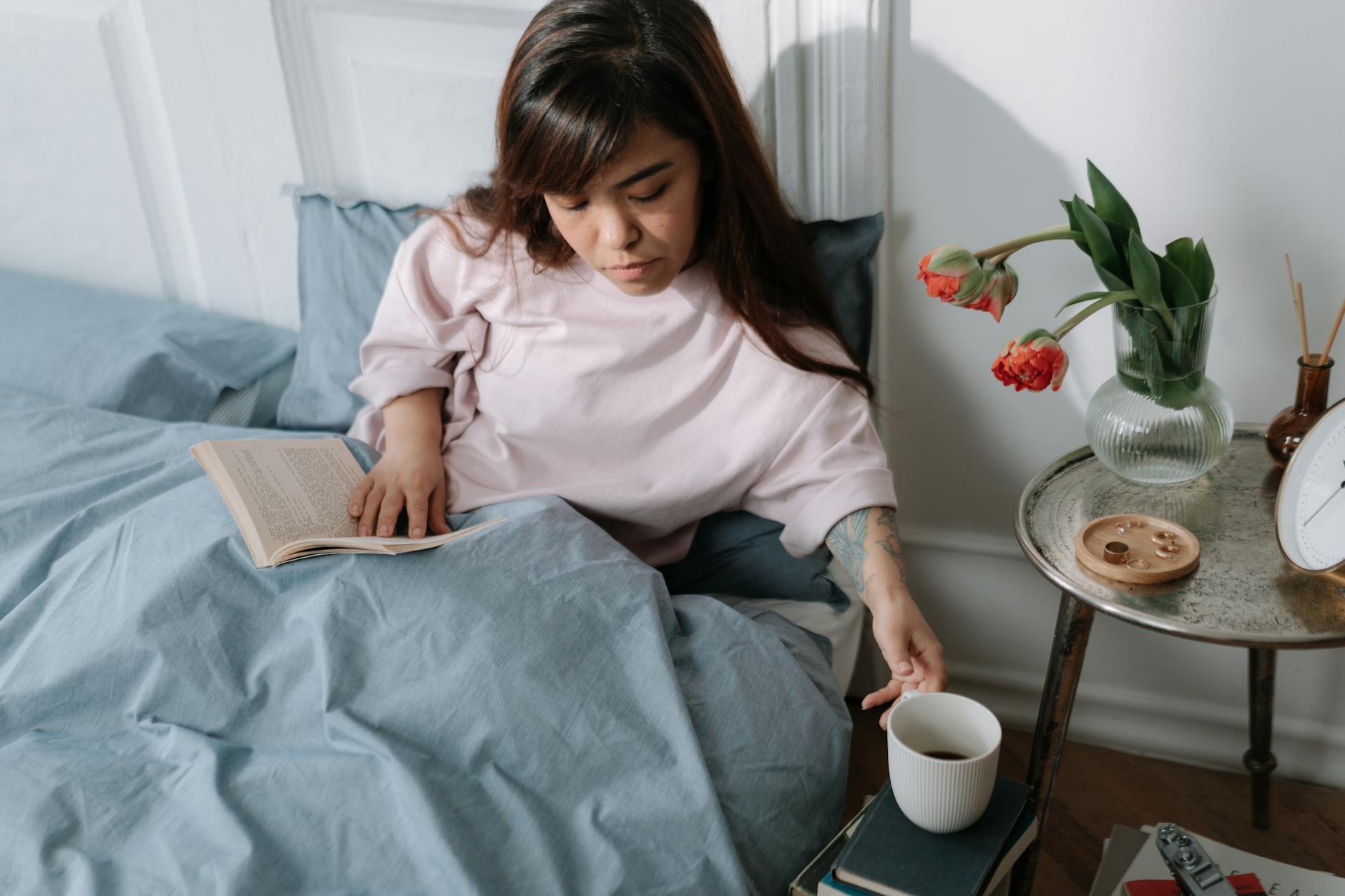 A woman resting in bed in soft morning light, reading and reaching for a warm mug on her bedside table — a calm sleep-and-wind-down scene for an article on ashwagandha and sleep quality