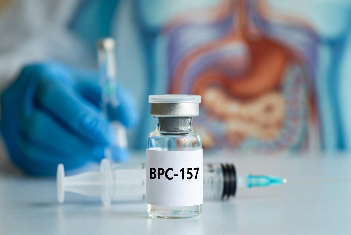 A clear glass research peptide vial labelled BPC-157 on a lab bench, a syringe behind it and a gloved researcher's hand near a blurred anatomical stomach and gut illustration