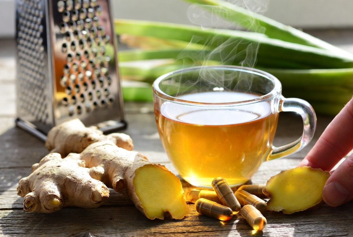 Fresh knobbly ginger root sliced open on a wooden table beside a steaming cup of golden ginger tea, amber ginger capsules, a metal grater and green ginger shoots, with a hand reaching toward the cup