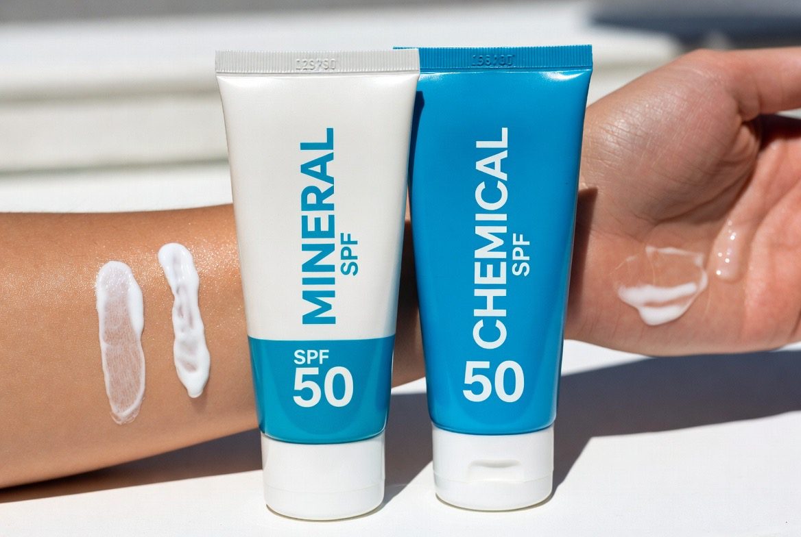 Two sunscreen tubes labeled MINERAL SPF 50 and CHEMICAL SPF 50 held against a bare forearm in bright light, with a thick white mineral swatch beside a sheer clear chemical swatch on the skin
