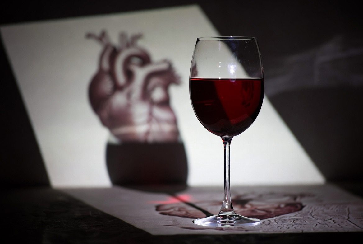 A single glass of red wine casting a long shadow in moody dramatic light, with an anatomical human heart illustration blurred behind it, evoking the collapsed heart-healthy moderate drinking myth
