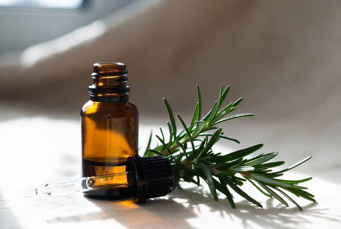 An amber glass dropper bottle of rosemary essential oil with a fresh green rosemary sprig beside it on a light surface in soft natural light
