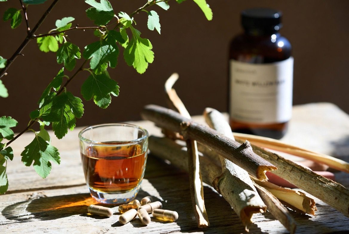 Curled strips of dried white willow bark beside a leafy Salix branch, herbal capsules, a small glass of amber willow bark tea, and an amber supplement bottle as a traditional natural pain remedy
